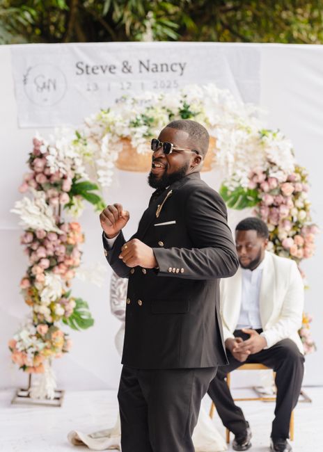 A man with dark skin, wearing sunglasses and a black suit with gold buttons, is smiling and holding a joyful pose outdoors during a wedding celebration. In the background, a seated man dressed in whit