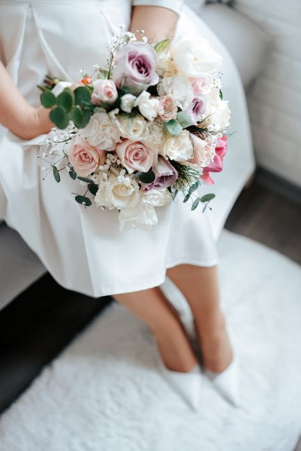 A person dressed in a white gown, sitting on a white cushioned surface, holding a large, lush bouquet of fresh flowers. The bouquet includes pale pink roses, white peonies, lavender lisianthus, and sm