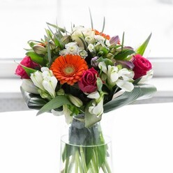 Showcasing the artistry and local flair of a skilled Stanmore florist, this exquisite floral arrangement marries vibrancy with grace. Four gerbera daisies-two a brilliant autumnal orange, two a delicate, creamy peach-radiate from the heart of the bouquet, each anchored by a textured, brown center. Deep magenta roses, their petals rich and tightly whorled, offer strength and drama, evoking the heritage and warm welcomes of Stanmore's close-knit community. Among these vivid highlights, clusters of pristine white alstroemeria and fine white spray roses are gently interspersed, lending layers of light and subtlety. Whisper-soft pale pink blossoms and minute white fillers emerge between glossy leaves, conjuring visions of wild blooms along Stanmore Common. The flowers are carefully nested in a lush ring of curved, broad aspidistra leaves that echo the natural abundance of local parks. Slender stems of vibrant greenery add depth and motion, creating a full, harmonious look that rises from the transparent vase where the green stems are clearly visible, speaking of garden freshness. Soft, ambient daylight enriches the floral colours against the pure backdrop, giving the scene both tranquility and unspoken joy. Perfect as a thoughtful gift, a table centrepiece, or a celebratory gesture, this bouquet imbues any Stanmore home with optimism and care.